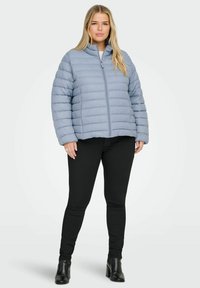 Světle modrá prošívaná bunda s vodorovným prošíváním, předním zipem a stojáčkovým límcem. Kombinováno s černými úzkými kalhotami a kotníkovými botami.