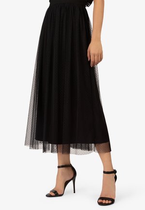 Zwarte midirok van tule met een stippenpatroon en een geplooide taille, gecombineerd met zwarte hoge sandalen met bandjes en een gespdetail.