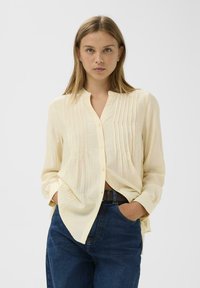 Lichtgele blouse met een knoopsluiting aan de voorkant, mandarin kraag en verticale plooien; gecombineerd met donkere blauwe jeans die een ontspannen pasvorm tonen.