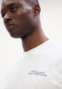 Tricou alb din bumbac cu guler rotund, având inscripționat cu fir albastru textul „fishing good MARC O POLO DENIM” pe partea stângă a pieptului.