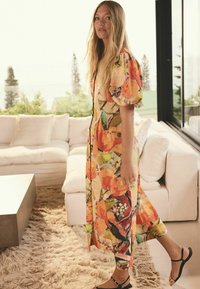 Kvetinové šaty v oranžových a zelených odtieňoch s nadýchanými rukávmi, po kolená, so zúženým pásom. Čierne sandále dotvárajú outfit.