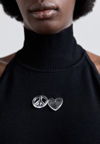 Des épingles en résine transparente en forme de symbole de la paix et de cœur, présentées sur un vêtement noir à col montant côtelé.