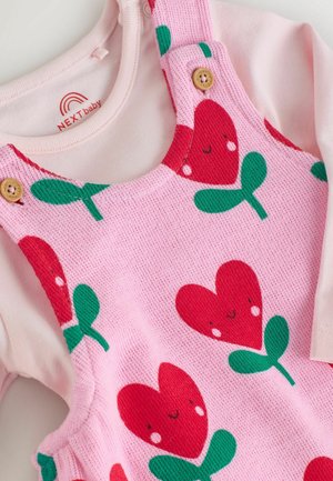 Růžové pletené šaty s overalovým střihem, zdobené vzorem srdce a listů, s dřevěnými knoflíky a měkkým béžovým topem s dlouhým rukávem pod nimi.