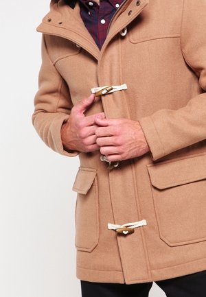 Homme boutonnant des boutons à bascule sur un manteau duffle couleur chameau avec de grandes poches à rabat, portant une chemise à carreaux foncée en dessous.