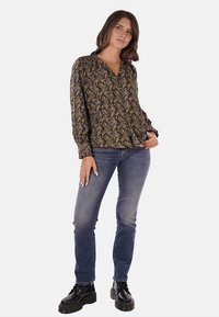 Blusa scura a fantasia con scollo a V, maniche lunghe e polsini elasticizzati, abbinata a jeans blu e stivali neri.