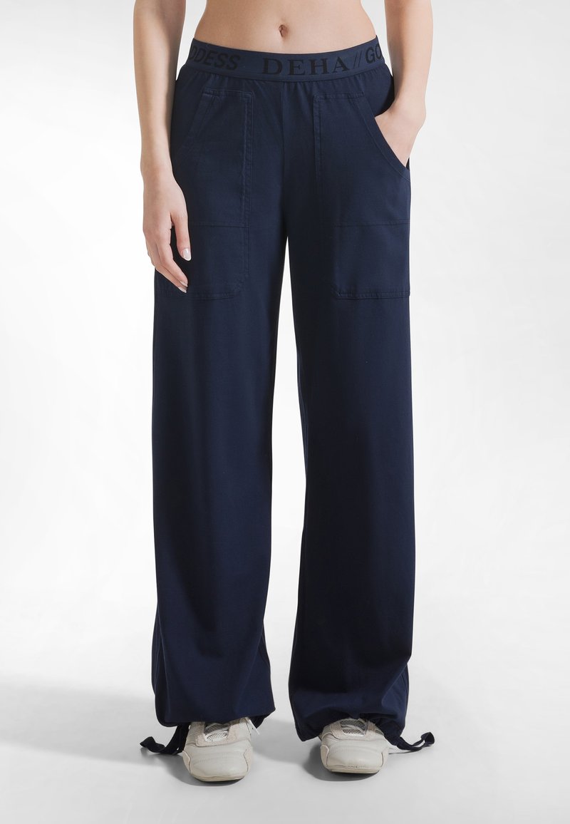 Granatowe spodnie o szerokich nogawkach z elastycznym pasem z czarnym logo, dwoma bocznymi kieszeniami i regulowanymi troczkami na dole nogawek. Gładka faktura tkaniny.