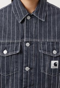 Donkerdenim jacket met witte streepjes, voorzien van zilveren drukknoopsluitingen, twee borstzakken en een zichtbaar logo-op-patch op de linkerzak.