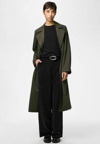 Lühikeste juustega noor inimene kannab oliivirohelist trentšmantlit, musta tuba, laiade säärega musti pükse vöökohaga ja musti kingi, seisab valgel taustal.