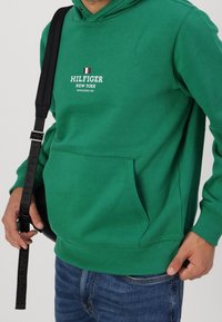 Grüne Kapuzenjacke mit einer Vorderentasche, die den Schriftzug "HILFIGER NEW YORK" und eine kleine italienische Flagge darüber zeigt. Das Material wirkt weich und strukturiert.
