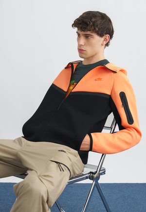 Młody mężczyzna siedzący na metalowym krześle, ubrany w pomarańczowo-czarną kurtkę zapinaną na zamek i beżowe spodnie typu cargo, patrzący na bok na tle gładkiej powierzchni.