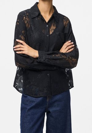 Kobieta w czarnej, prześwitującej koronkowej koszuli na guziki z długim rękawem, z założonymi rękami i ciemnoniebieskich jeansach na jednolitym tle.