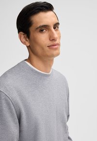 Szary sweatshirt z okrągłym dekoltem i ściągaczami, wykonany z miękkiego materiału. Model ma ciemne włosy i stoi pod lekkim kątem.