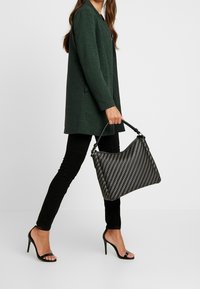Sac tissé noir à finition texturée, doté d'une forme structurée et d'une anse en cuir, assorti à un manteau vert et un pantalon noir.