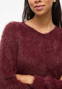 Cardigan bordeaux profond avec une texture duveteuse, décolleté en V et fermeture à boutons. Il possède des manches longues douces avec une coupe ample et un léger lustre.
