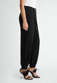 Pantaloni neri su misura con pieghe, caratterizzati da motivi decorativi. Abbinati a tacchi neri decorati con piume e cinture sottili.