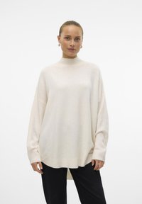 Oversized cream sweater with a high neck, ribbed cuffs, and side slits. Made from soft fabric, offering a relaxed fit and minimal texture.