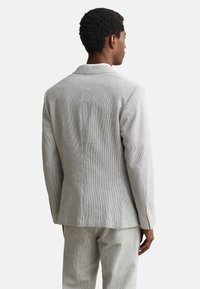 Homme de dos portant une veste de costume gris clair texturée et un pantalon assorti sur un fond blanc uni.