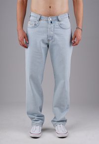 Mann in hellblauen geraden Jeans und weißen Turnschuhen, der vor einem schlichten grauen Hintergrund steht, mit entspannten Händen an den Seiten.