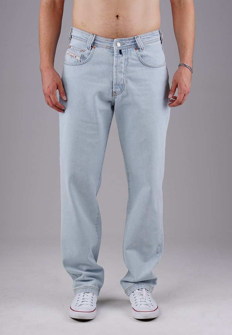 Mann in hellblauen geraden Jeans und weißen Turnschuhen, der vor einem schlichten grauen Hintergrund steht, mit entspannten Händen an den Seiten.