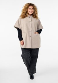 Cappotto in misto lana beige con colletto alto, chiusura a bottoni e due tasche frontali. Indossato sopra un top nero a maniche lunghe e abbinato a jeans scuri.
