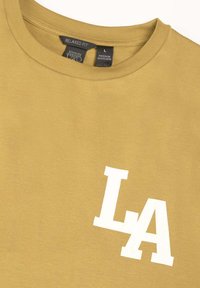 T-shirt en coton jaune avec un design décontracté, un col rond et un logo "LA" blanc imprimé sur le devant. Étiquette de taille L visible.