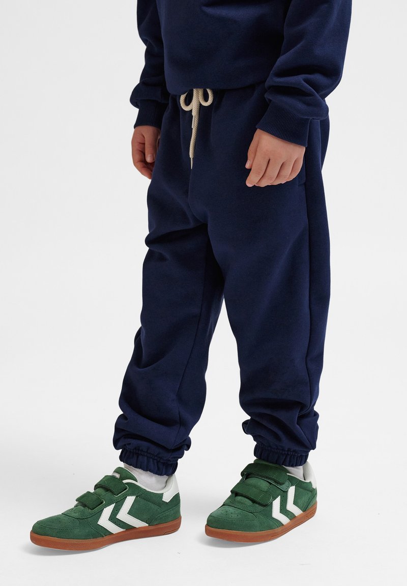 Pantaloni della tuta navy con polsini elasticizzati, abbinati a sneakers verdi con accenti bianchi e cinturini in Velcro. Tessuto morbido.