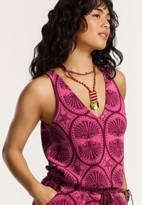 Rózsaszín, ujjatlan blúz kagylómintával, sötét tónusokkal kiemelve, többrétegű gyöngy nyakláncokkal kiegészítve. Az anyag lágy és textúrált hatású.