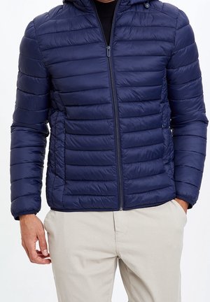 Mann trägt eine marineblaue gesteppte Jacke mit Frontreißverschluss und beige Hosen, Hände in den Taschen, vor weißem Hintergrund.