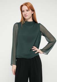 Dark green blouse with sheer long sleeves, high neckline, and a flowing, textured fabric. Paired with loose black trousers.