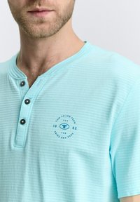 Šviesiai mėlynas trumpomis rankovėmis henley marškinėliai su tekstūruota dryžuota raštu, trimis juodais mygtukais ir apvaliu mėlynu logotipu ant krūtinės.