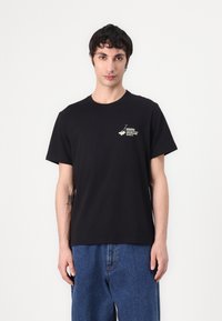 Black cotton t-shirt with short sleeves, featuring a small graphic design on the left chest. Smooth texture; classic fit.