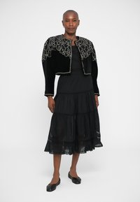 Black velvet cropped jacket with intricate cream embroidery, paired with a black tiered dress. Smooth texture and lightweight design.