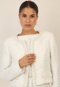 White, textured blazer with pearl accents along the collar and front. Features a subtle shimmer and fitted design, with a side pocket.
