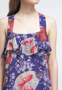 Ärmlös lila klänning med breda band, blommönster och röda fiskar, samt en volangdetalj vid halslinningen på en ljus bakgrund.