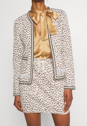 Vrouw draagt een wit-bruin patroon jas- en rokkenset met zwarte biezen, gecombineerd met een gouden satijnen blouse met grote strik bij de hals.