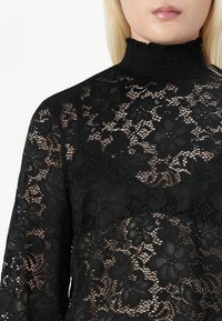 Črna čipkasta turtlenecka majica z cvetličnimi vzorci, teksturiranimi rokavi in smokanjem ob vratu, zasnovana za prilegajočo silhueto.