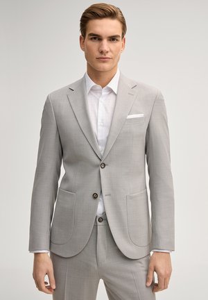 Giovane uomo con completo grigio chiaro, camicia bianca e fazzoletto da taschino, in piedi davanti a uno sfondo chiaro e uniforme.