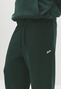 Pantaloni de trening verde închis, confecționați dintr-un material moale. Au o talie elastică cu șnur și un mic logo alb pe piciorul inferior.