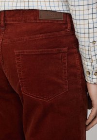 Cordhosen in tiefrot mit strukturierter Oberfläche, ausgestattet mit einer Gesäßtasche und einem Leder-Brandlabel in der Nähe des Bundes.