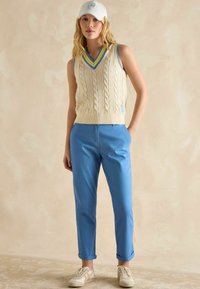Kremowy sweter bez rękawów w warkoczowy splot z niebieskimi i żółtymi paskami przy dekolcie w kształcie litery V, w zestawie z jasnoniebieskimi spodniami i beżowymi trampkami. W komplecie biały kapelusz.