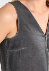 Top grigio senza maniche con zip frontale indossato da una persona, che mostra la parte superiore del torso e dell'area della spalla.