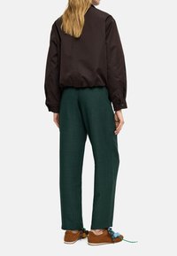 Brązowa kurtka bomberka o skróconym kroju, zestawiona z zielonymi, szerokimi spodniami o fakturze oraz brązowymi butami z kolorowymi sznurowadłami i pomponami.