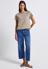 T-shirt beige à manches courtes avec un col rond, associé à un jean large bleu clair et des sandales à lanières argentées.