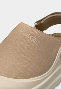 Κοντινό πλάνο μπεζ παπουτσιού χωρίς κορδόνια με λογότυπο UGG, υφασμάτινο επάνω μέρος με υφή και άκρη λευκής καουτσούκ σόλας.