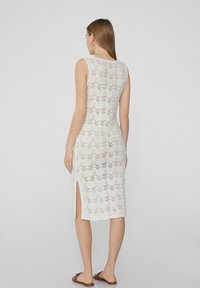 Weißes Strickkleid mit tailliertem Schnitt, kompliziertem Spitzenmuster und seitlichen Schlitzen für einen angenehmen Bewegungsspielraum. Kombiniert mit flachen braunen Sandalen.