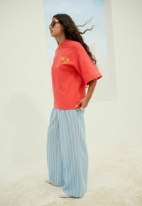 Korálové oversized tričko s potiskem "PALM" v žluté barvě, v kombinaci s modrými pruhovanými širokými kalhotami. Model má na sobě černé sluneční brýle.
