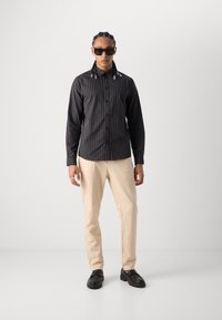 Schwarz gestreiftes Hemd mit Kragen, beige Hose und schwarze Schuhe. Das Hemd hat vertikale Streifen und Schneiderakzente.