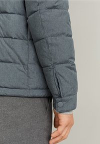 Chaqueta de plumas gris con un patrón cosido, cierre de botón en la manga y una textura suave, con un puño de punto gris liso por debajo.