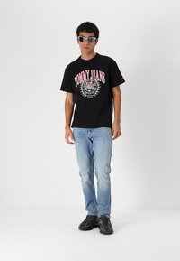 Tricou din bumbac negru cu imprimeu roz "TOMMY JEANS", asociat cu blugi decolorati și pantofi sport negri. Accesoriile includ ochelari de soare.