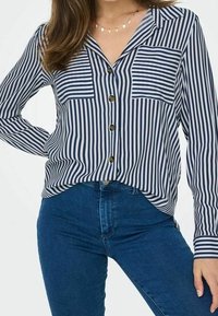 Camicia a righe verticali blu navy e bianche con maniche lunghe, due tasche sul petto e bottoni marroni, abbinata a jeans blu.